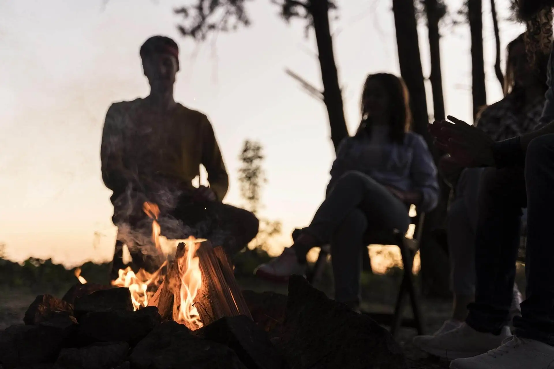Eine Gruppe von Freunden sitzt im Kreis um ein Lagerfeuer in der Abenddämmerung. Die lodernden Flammen und aufsteigender Rauch erhellen die Szene, während die Silhouetten der Menschen im Hintergrund vor dem sanften Licht des Sonnenuntergangs sichtbar sind. Einige der Freunde sitzen entspannt auf Campingstühlen, während eine Person ihre Hände aneinander reibt, als würde sie sich wärmen. Die Umgebung ist von Bäumen umgeben, was auf ein Outdoor- oder Camping-Abenteuer hindeutet. Die Atmosphäre wirkt gemütlich, entspannt und naturverbunden.