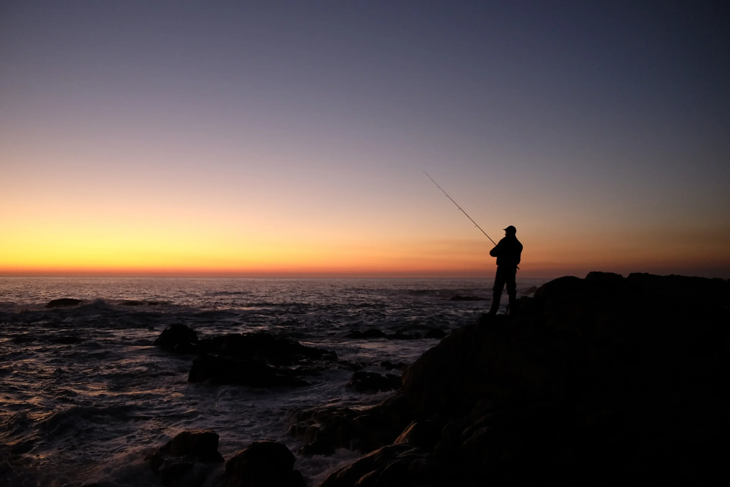 Warum Nachtangeln so besonders ist – Fische, Natur und die richtige Vorbereitung 22 Silhouette eines Mannes, der in der Dämmerung auf den Felsen steht und auf das Meer blickt, während er angelt.