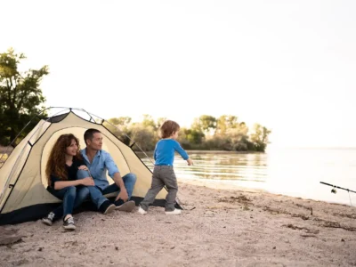 Eine Familie campt am Seeufer. Ein Mann und eine Frau sitzen entspannt vor einem Zelt, während ihr kleines Kind in einem blauen Pullover am Wasser spielt. Eine Angelrute liegt am Sandstrand. Die Szene vermittelt eine ruhige und glückliche Atmosphäre in der Natur.