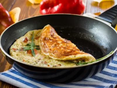 Omelette in einer schwarzen Pfanne mit goldbrauner Kruste, gefüllt mit Tomatenstückchen und Kräutern, garniert mit Rucola. Die Pfanne steht auf einem blau-weiß gestreiften Tuch, im Hintergrund liegen rote Paprika und Herbstblätter auf einem Holztisch.