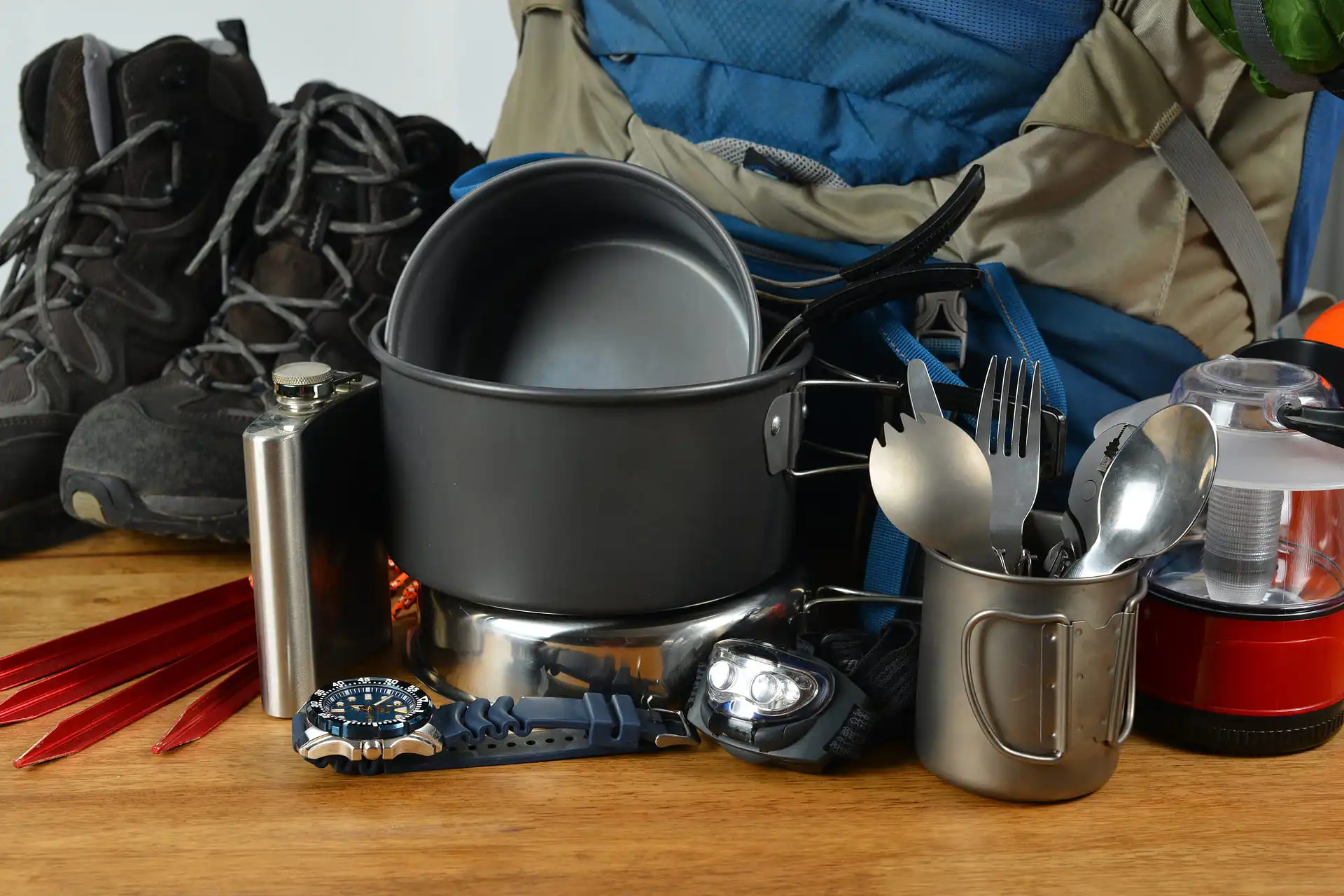 Campingausrüstung mit Fokus auf die Campingküche: Ein Campingkochtopf, Becher mit Besteck, eine Stirnlampe, eine Armbanduhr, eine Feldflasche, Zelt-Heringe und Wanderschuhe vor einem großen Rucksack auf einem Holztisch.
