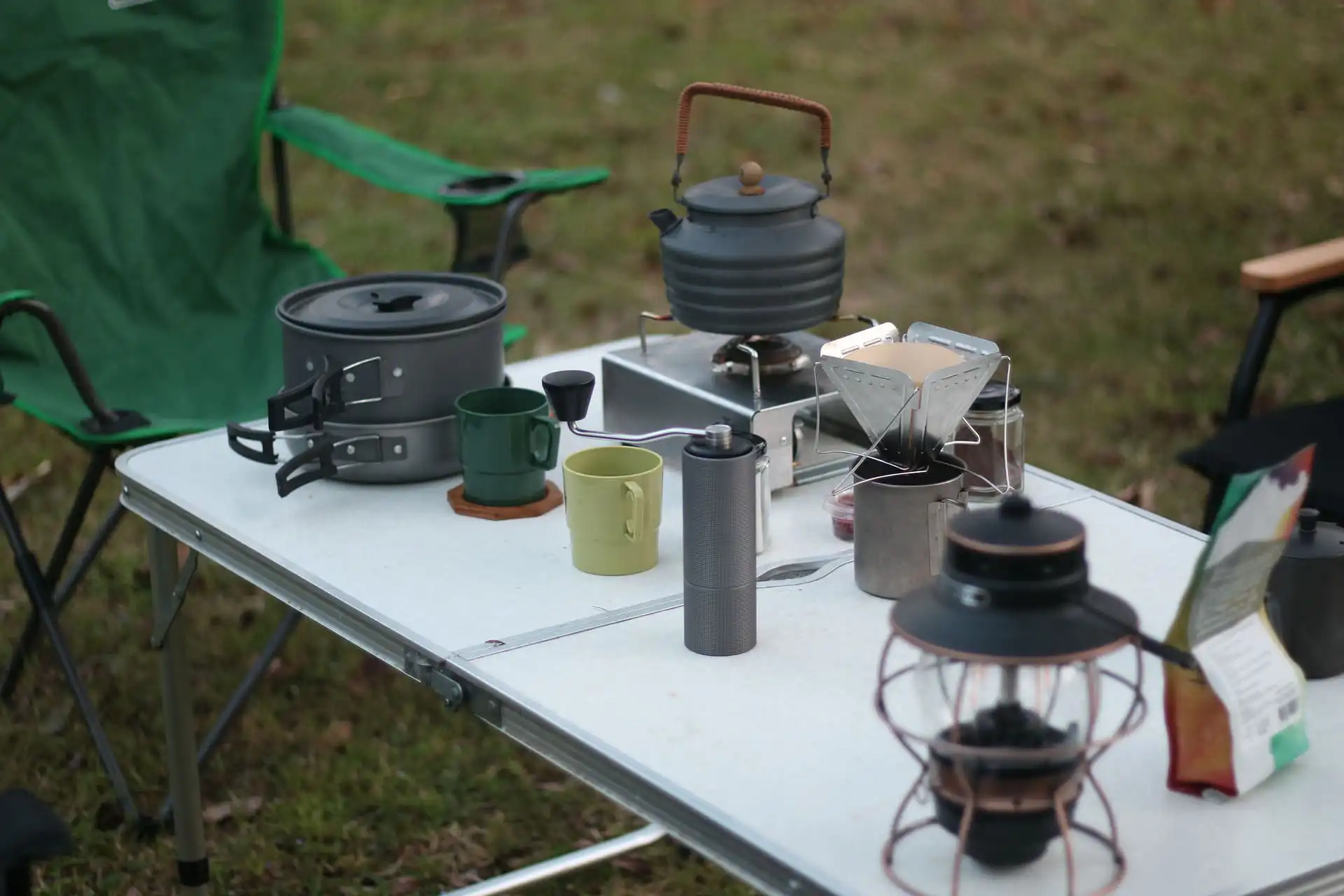 Campingküche auf einem Klapptisch im Freien: Campingkocher mit Teekessel, Stapel von Töpfen, Kaffeefilterhalter, Handkaffeemühle, Tassen, Laterne und weitere Utensilien. Im Hintergrund Campingstühle auf einer Wiese.
