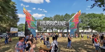 Eingangsbereich des Hurricane Festivals mit großem Schild und bunten Stoffbahnen an den Seiten. Zahlreiche Festivalbesucher strömen bei sonnigem Wetter durch das Tor auf das Gelände. Im Hintergrund sind Bäume, Zäune und Stände zu sehen, die das lebendige Festivalgeschehen einrahmen.