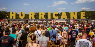Große Menschenmenge beim Einlass zum Hurricane Festival an einem sonnigen Tag, im Hintergrund große gelbe Buchstaben mit dem Schriftzug 'HURRICANE'.