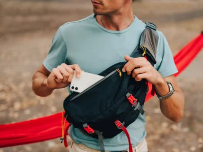 Ein junger Mann in einem hellblauen T-Shirt steckt ein Smartphone in die Vordertasche seiner schwarzen Umhängetasche mit roten Details. Im Hintergrund ist eine rote Hängematte zu sehen, der Boden ist mit trockenen Blättern bedeckt.