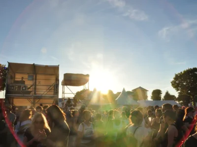 Menschenmenge auf einem Open-Air-Festival bei Sonnenuntergang. Im Hintergrund sind Bühnenaufbauten und Pavillons zu sehen, aber keine Band oder Musiker auf der Bühne – nur ein DJ im Technikbereich sichtbar. Die Lichtstimmung wirkt warm und sommerlich, mit auffälligen Sonnenstrahlen und Lens Flare im Bild.