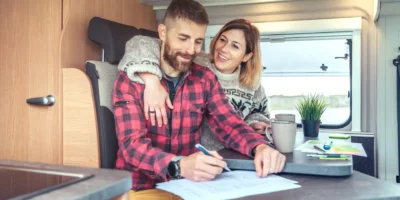 Ein fröhliches Paar sitzt im Wohnmobil am Tisch, der Mann unterschreibt Dokumente – vermutlich Kauf- oder Mietunterlagen für das Wohnmobil. Die Frau umarmt ihn lächelnd von hinten. Auf dem Tisch liegen Tassen, ein Kugelschreiber, eine Zimmerpflanze und Kartenmaterial.