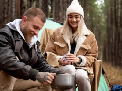 Ein bärtiger Mann mit Tattoos an den Armen bereitet beim Wintercamping in einem Waldstück Essen in einem Topf zu, während eine junge Frau mit blonder Haaren, die eine weiße Mütze und eine dicke beige Jacke trägt, lächelnd danebensitzt und eine Tasse in den Händen hält. Im Hintergrund ist ein Zelt und ein dichter Wald zu sehen. Die Szene vermittelt Gemütlichkeit und die Nutzung der Restwärmemethode zum Kochen im Freien.