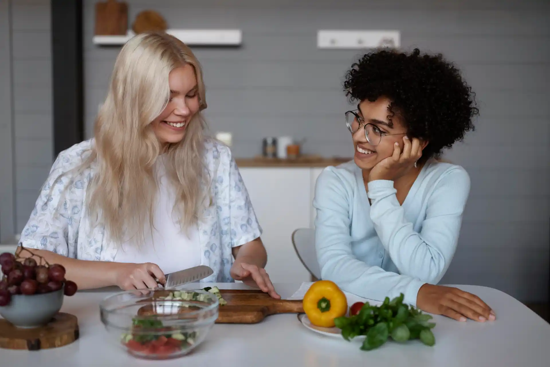 Zwei Frauen sitzen lächelnd in einer modernen Küche, während eine von ihnen Zutaten für einen Linsensalat auf einem Holzbrett schneidet. Auf dem Tisch liegen frisches Gemüse wie gelbe Paprika, Basilikum, Trauben und eine Glasschüssel mit Tomaten und Kräutern. Die Stimmung ist entspannt und harmonisch.