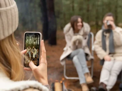 Drei junge Frauen genießen einen Moment beim Camping im Wald. Eine der drei Frauen steht im Vordergrund und mit einem Blick über ihre Schulter kann man sehen, dass sie gerade ein Foto von ihren zwei Freundinnen macht. Sie tragen warme Winterkleidung mit Fellbesatz und dicken Schals, was auf kühle Temperaturen hinweist – ein Hinweis auf den Trend zum Ganzjahres-Camping. Eine der zwei Frauen trinkt aus einem Thermobecher während neben ihren Füßen ein Lagerfeuer brennt. Sie sitzen auf Klappstühlen, während sie fotografiert werden. Die Szene vermittelt Geselligkeit, Naturverbundenheit und Komfort – typische Elemente der Camping Trends 2025, bei denen Erlebnisse mit Freunden, moderne Ausrüstung und stylische Funktionalität im Vordergrund stehen.