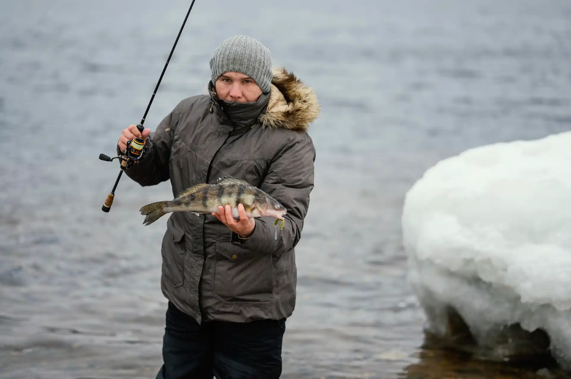Angler im Wintermantel hält frisch gefangenen Barsch am verschneiten Seeufer – Angeln im Winter