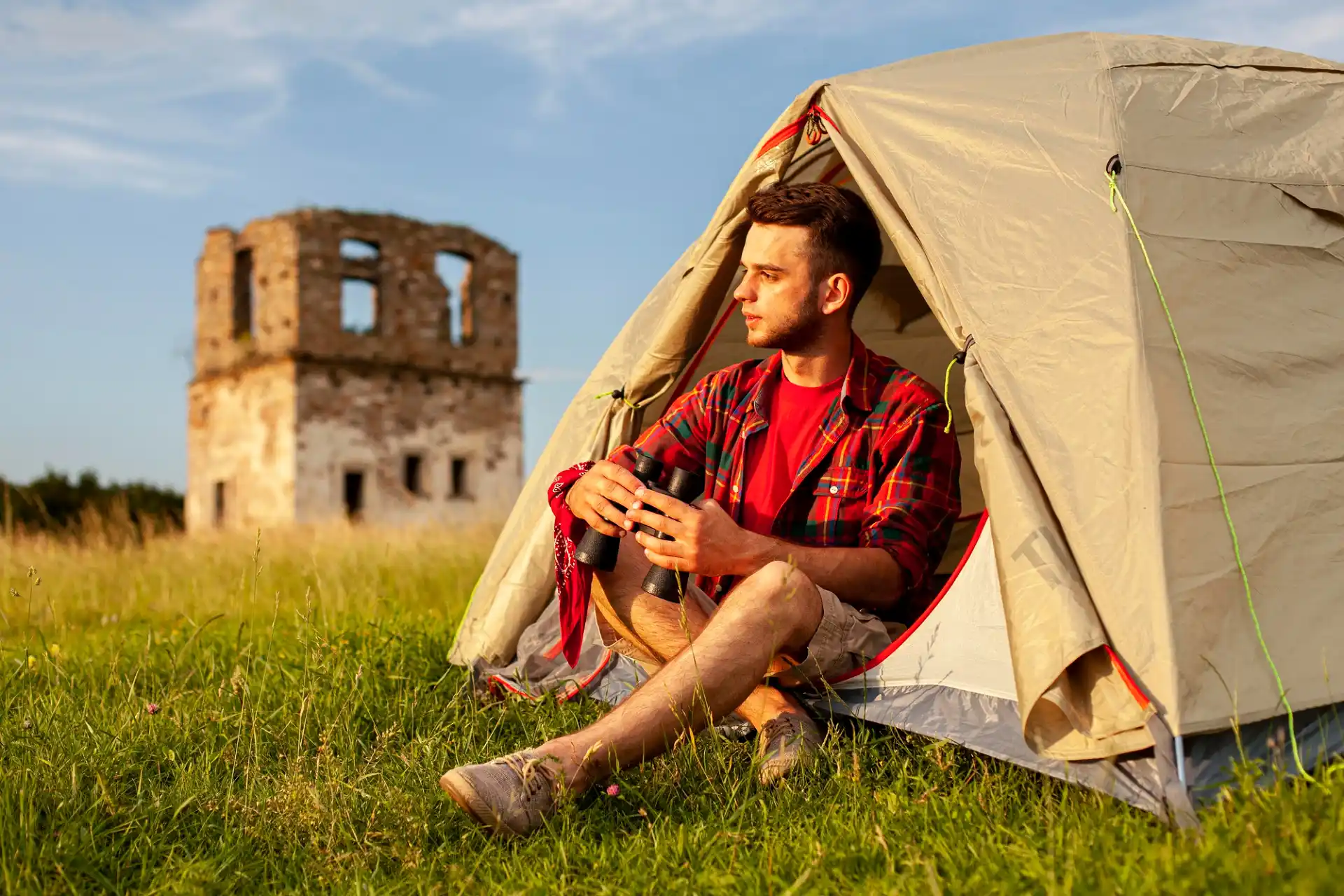 Ein junger Mann sitzt im Eingangsbereich eines hellen Zeltes auf einer grünen Wiese und blickt nachdenklich in die Ferne, während er ein Fernglas in den Händen hält. Er trägt ein rot-kariertes Hemd über einem roten T-Shirt und kurze Hosen. Im Hintergrund ist eine alte, verfallene Steinruine unter einem klaren blauen Himmel zu sehen. Die Szene vermittelt eine ruhige und naturnahe Atmosphäre, die ideal für Camping in Frankreich ist.