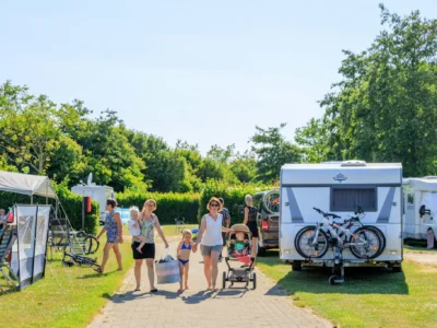 Ein Campingplatz liegt direkt hinter den Dünen an der Nordsee. Familien sitzen entspannt unter der Markise ihres Wohnmobils, während Kinder auf der Wiese Fußball spielen. Im Hintergrund sieht man mehrere Zelte, die Dünen und das Meer mit einem Segelboot am Horizont. Das Bild wirkt authentisch, lebendig und spiegelt die typische Atmosphäre eines Küstencampings wider.
