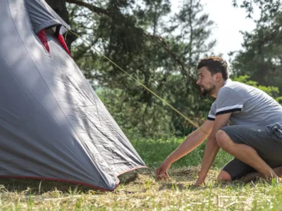 Ein Mann mit kurzem Bart und in Freizeitkleidung (graues T-Shirt und Shorts) kniet auf dem Gras und ist dabei, ein graues Zelt auf einem Waldcampingplatz aufzubauen. Er fasst mit einer Hand an den unteren Rand des Zeltes, während die andere Hand den Boden berührt. Eine gelbe Abspannleine ist gespannt. Der Fokus liegt auf der Anstrengung und Konzentration des Mannes beim Zelt aufbauen inmitten der Natur.
