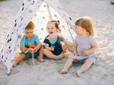Ein warmes Sommerfoto zeigt drei Kleinkinder, die vergnügt im Sand sitzen und spielen. Sie befinden sich unter einem kleinen, dreieckigen Spielzelt mit einem verspielten Muster und sind von Sand umgeben. Das mittlere Kind lacht ausgelassen und hält eine Schaufel in der Hand, während die beiden anderen Kinder mit Sandspielzeug beschäftigt sind. Die Szene fängt die Freude und das einfache Vergnügen des Camping am Meer mit Kindern ein.