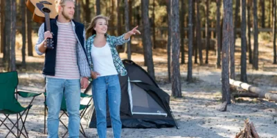 Ein helles, horizontales Foto zeigt ein lächelndes junges Paar, das vor seinem kleinen, dunklen Zelt auf einem sandigen Waldboden steht. Der Mann mit Gitarre auf der Schulter und die Frau, die in die Ferne zeigt, wirken entspannt und abenteuerlustig. Im Vordergrund sind ein Campingstuhl und ein kleines Lagerfeuer aus Holzscheiten zu sehen. Die Szene vermittelt eine naturnahe Campingatmosphäre in einem Waldgebiet, wie es für einige Campingplätze in den Niederlanden (z.B. in der Veluwe) typisch ist.