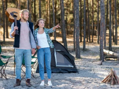 Ein helles, horizontales Foto zeigt ein lächelndes junges Paar, das vor seinem kleinen, dunklen Zelt auf einem sandigen Waldboden steht. Der Mann mit Gitarre auf der Schulter und die Frau, die in die Ferne zeigt, wirken entspannt und abenteuerlustig. Im Vordergrund sind ein Campingstuhl und ein kleines Lagerfeuer aus Holzscheiten zu sehen. Die Szene vermittelt eine naturnahe Campingatmosphäre in einem Waldgebiet, wie es für einige Campingplätze in den Niederlanden (z.B. in der Veluwe) typisch ist.