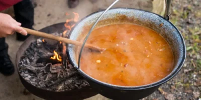 Ein Detailfoto aus der Vogelperspektive zeigt einen großen, schwarzen Emaille-Kessel, der über einem offenen Feuer in einer runden Feuerschale hängt. Der Kessel ist mit einem dampfenden, orange-braunen Eintopf gefüllt, der mit einem langen Holzlöffel von einer Person umgerührt wird, deren Hand am linken Bildrand sichtbar ist. Die Szene, die sich im Freien abspielt, demonstriert die rustikale Zubereitung eines herzhaften One Pot Gerichtes über dem Lagerfeuer.