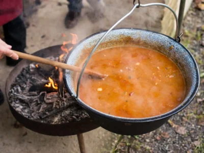 Ein Detailfoto aus der Vogelperspektive zeigt einen großen, schwarzen Emaille-Kessel, der über einem offenen Feuer in einer runden Feuerschale hängt. Der Kessel ist mit einem dampfenden, orange-braunen Eintopf gefüllt, der mit einem langen Holzlöffel von einer Person umgerührt wird, deren Hand am linken Bildrand sichtbar ist. Die Szene, die sich im Freien abspielt, demonstriert die rustikale Zubereitung eines herzhaften One Pot Gerichtes über dem Lagerfeuer.