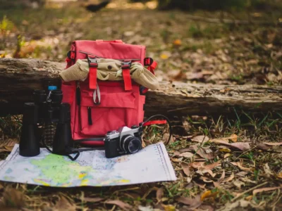 Ein roter Wanderrucksack liegt vor einem umgestürzten Baumstamm auf dem Boden eines Waldes, der mit braunen Blättern bedeckt ist. Im Vordergrund sind eine Landkarte, ein Fernglas und eine analoge Kamera ausgebreitet. Das Bild ist im Stil einer Stillleben-Aufnahme gehalten und zeigt die essenzielle Outdoor Ausrüstung für eine Wanderung oder einen Campingausflug.